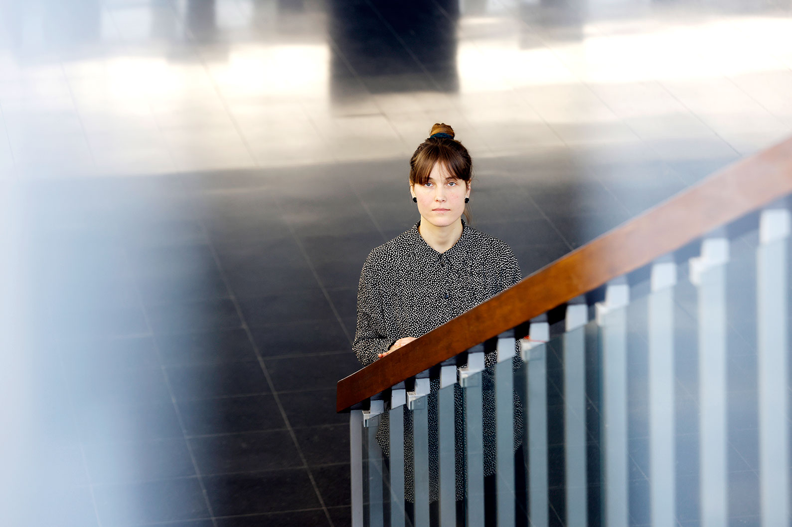 Kuvassa haastateltava Tiina Järvi seisoo Tampereen yliopiston aulassa portaikon kaiteen reunan vieressä ja katsoo kameraan. 