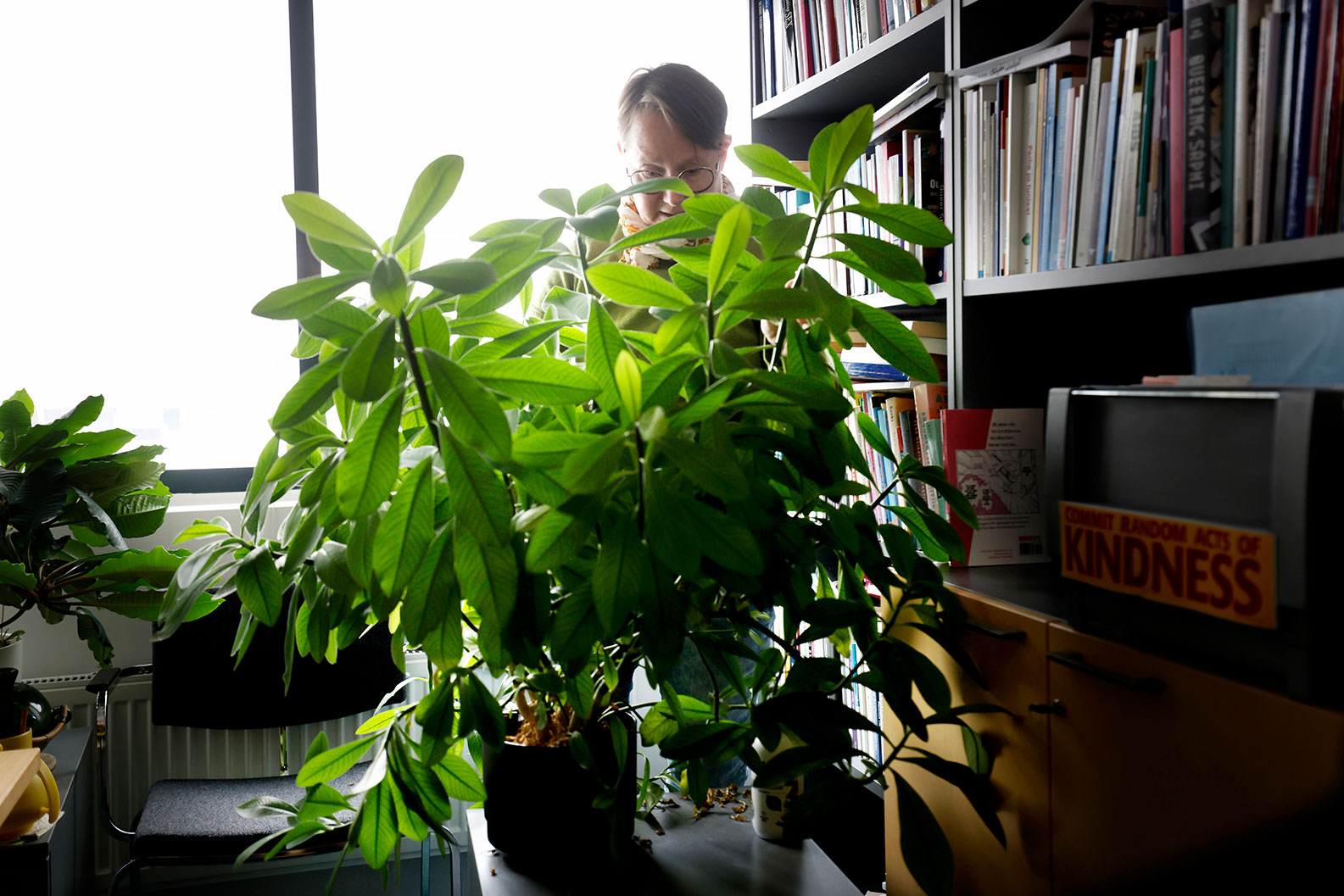 Tuula Juvonen seisoo työhuoneessaan kirjahyllyn vieressä ja suuren viherkasvin takana.