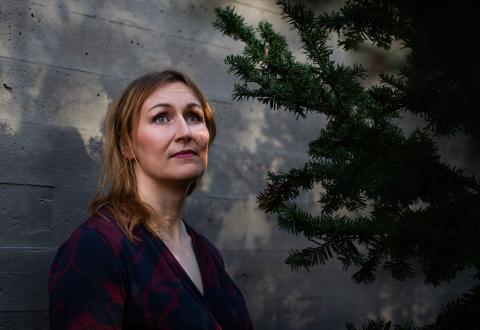 Kuvassa haastateltava Tiina Raevaara seisoo valkoista seinää vasten puun vieressä.